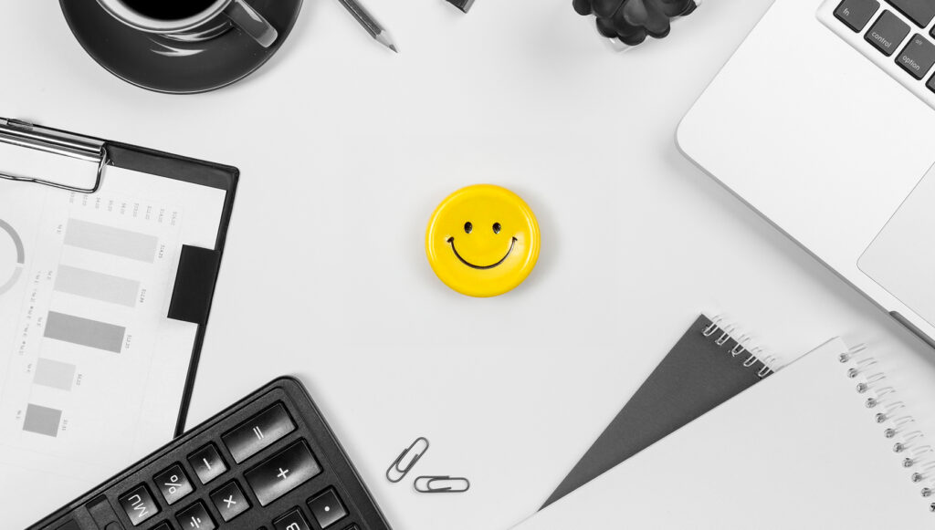 Overview Of A Desk With Coffee And Computers Surrounding A Bright Yellow Smiley Face Pin