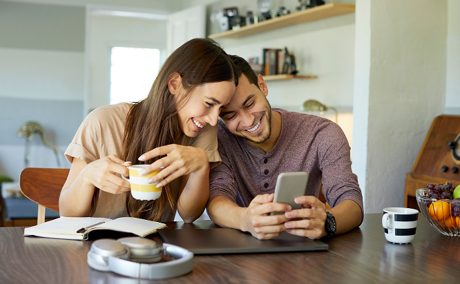 Young Couple At A Table Looking At Their Phone Smiling And Drinking Coffee